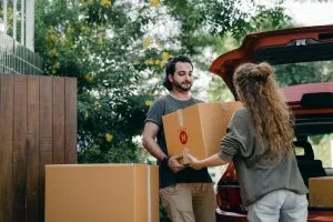 a man and a woman unpacking the car and carrying a moving box as they want to choose the best neighborhood.