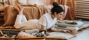 a woman lying on the floor between two suitcases full of clothes reading a step-by-step unpacking guide